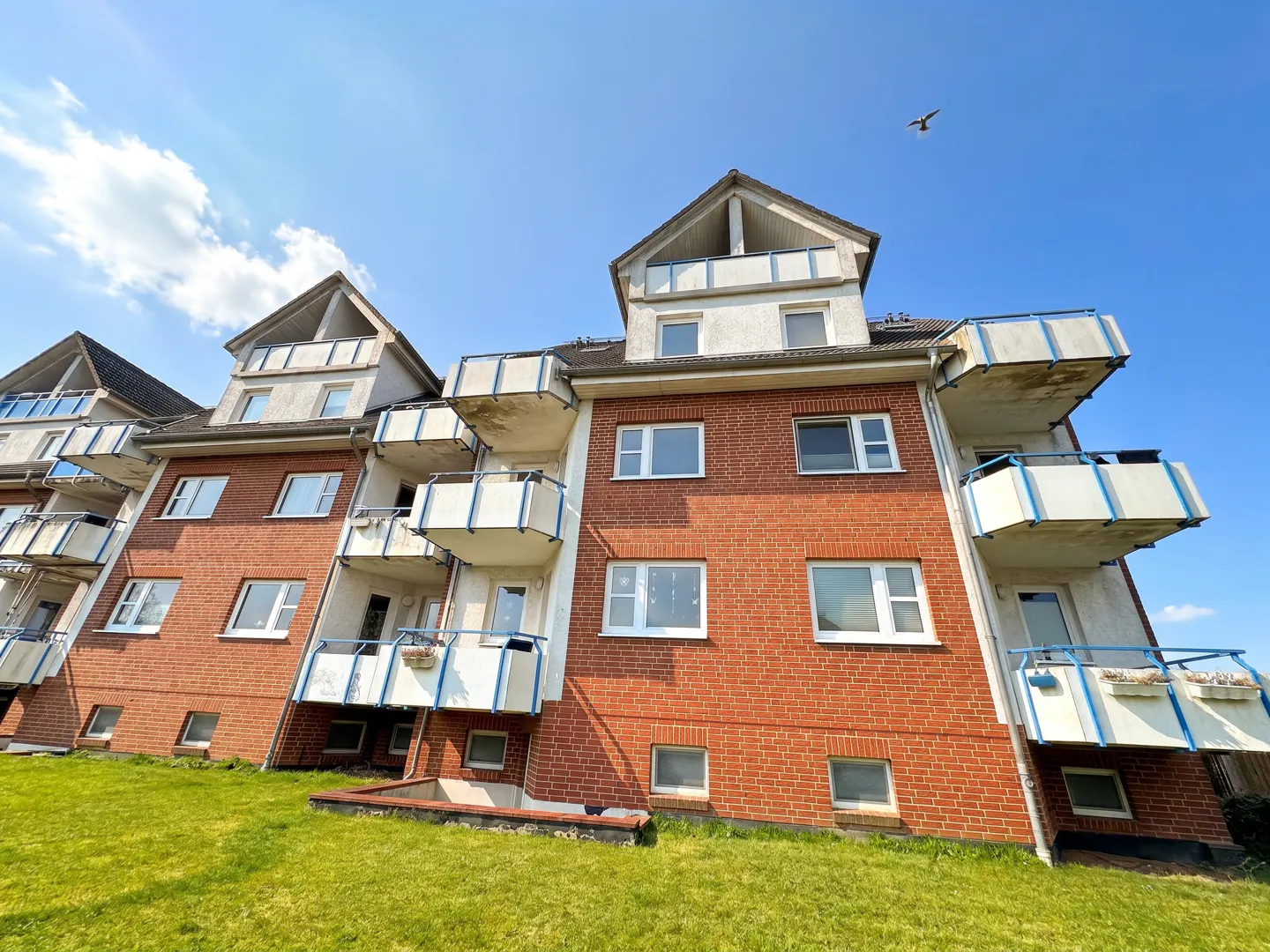 Exterior view of a red brick apartment building with white balconies under a blue sky with a bird flying above.