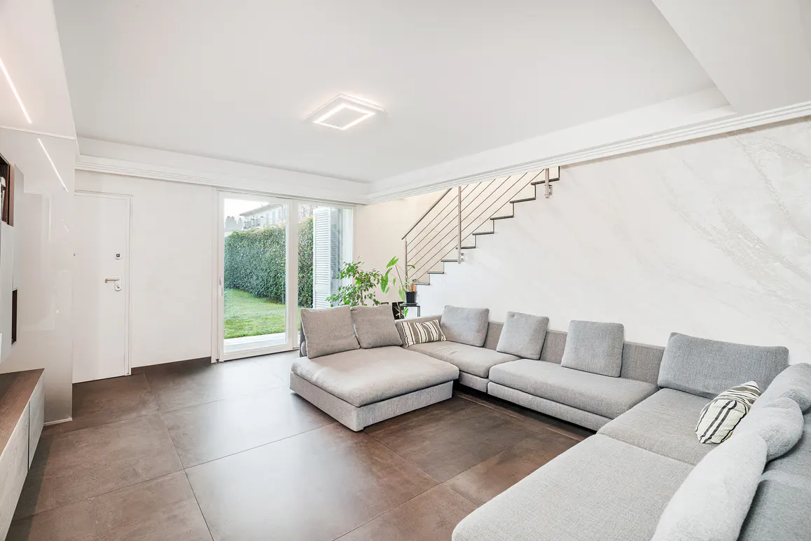 Bright living room with a gray sectional sofa, brown tile floor, and stairs with metal railing. Sliding glass doors lead to a green lawn.