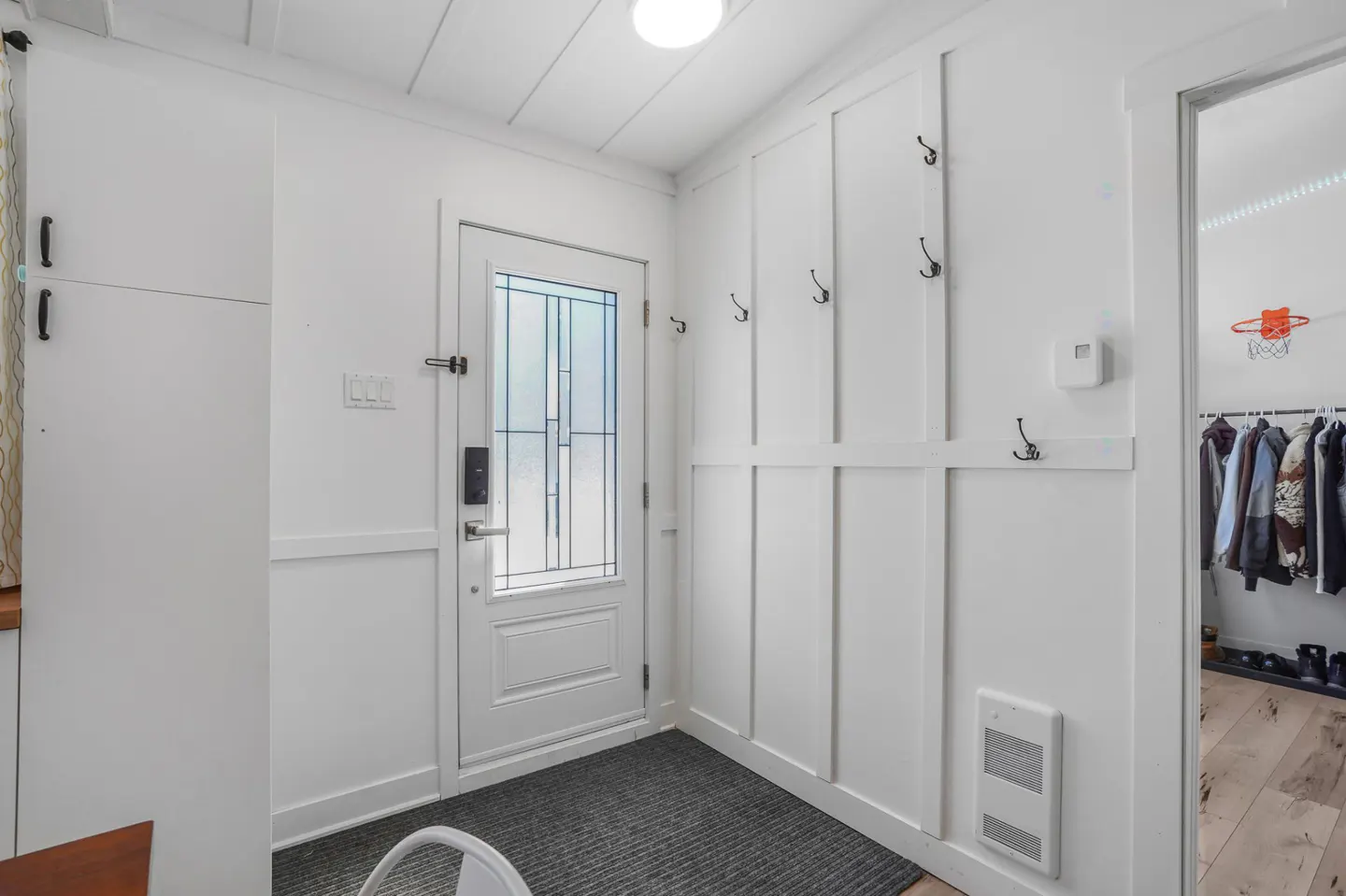Bright entryway with white walls, door with glass panels, and coat hooks. A gray rug covers the floor. A closet is visible through an open doorway.