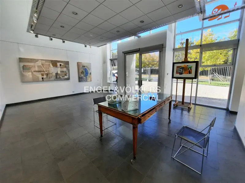 Bright gallery space with art on walls, a glass-topped table, chairs, and an easel with a painting near large windows.