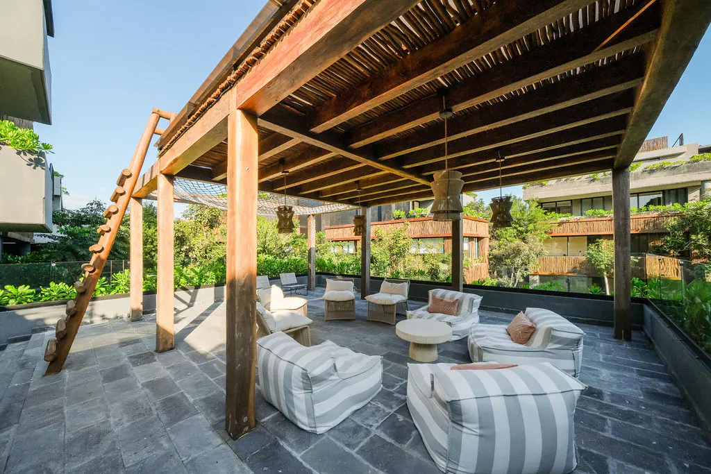 Outdoor patio with a wooden pergola, striped lounge chairs, and a small table on a stone floor surrounded by lush greenery.
