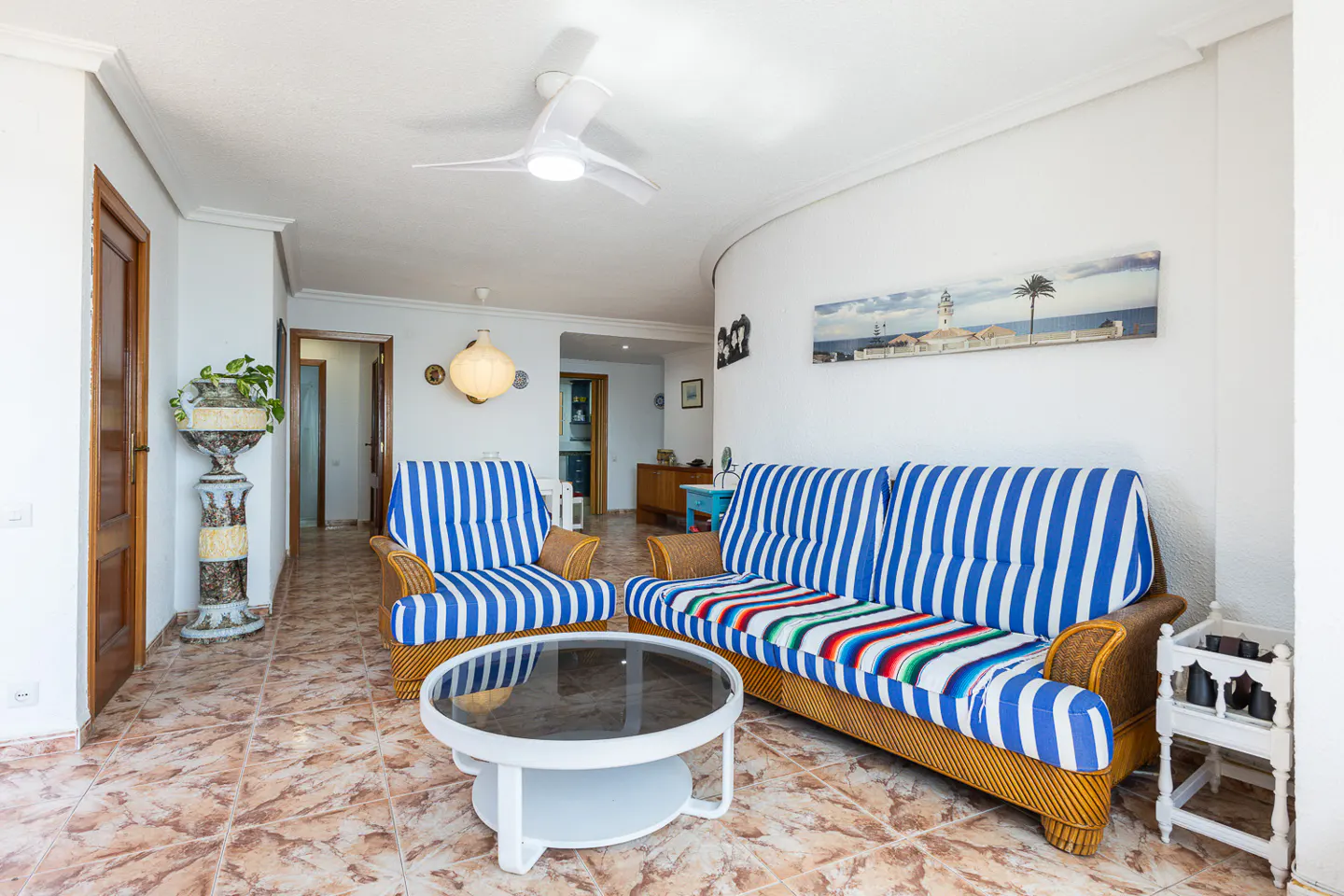 Bright living room with blue and white striped sofa and armchair, round white table, and tiled floor. A ceiling fan and coastal art add to the decor.