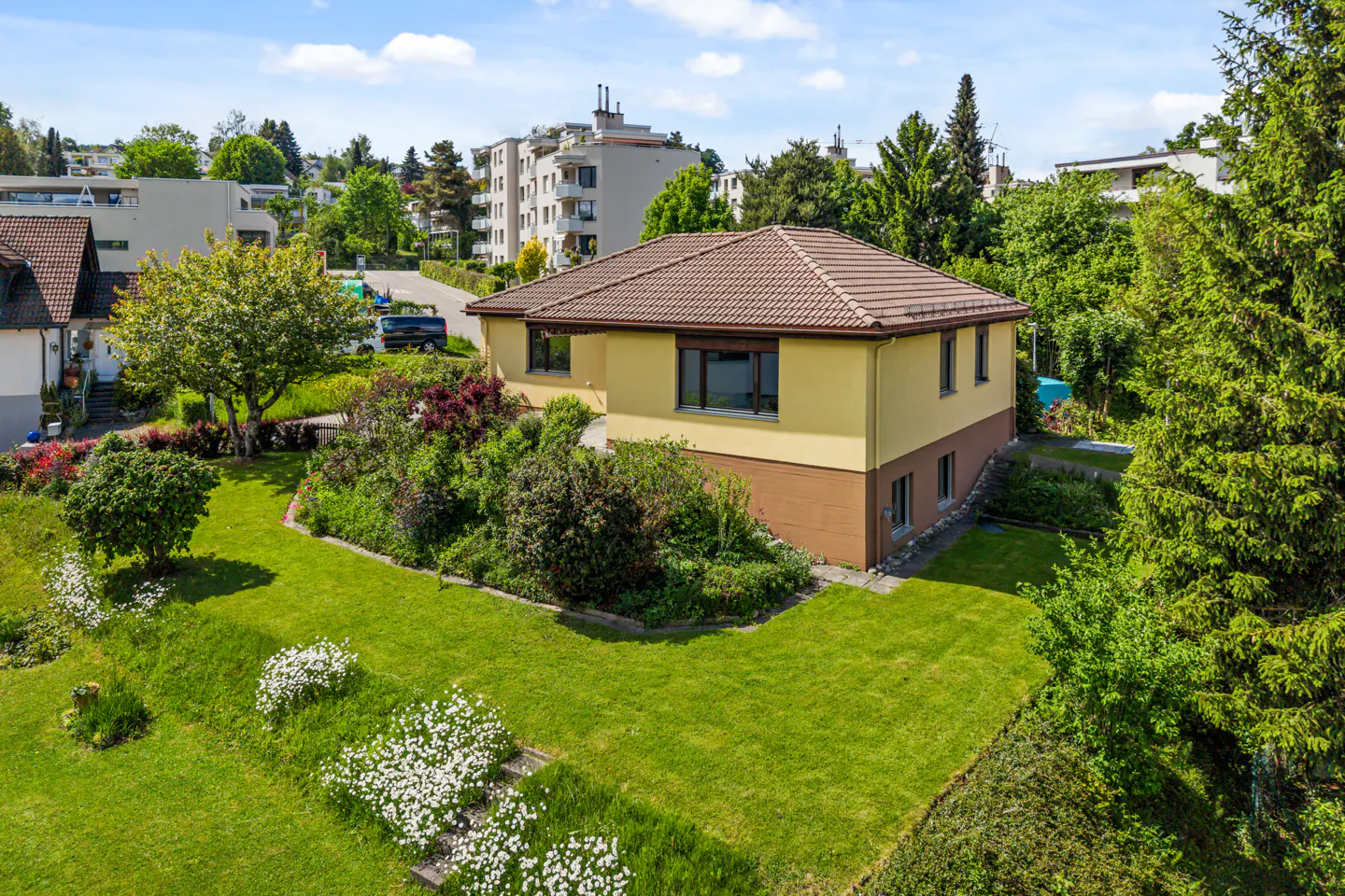 Two-story yellow house with a brown roof and trim, surrounded by lush green lawn, trees, and colorful flowerbeds.