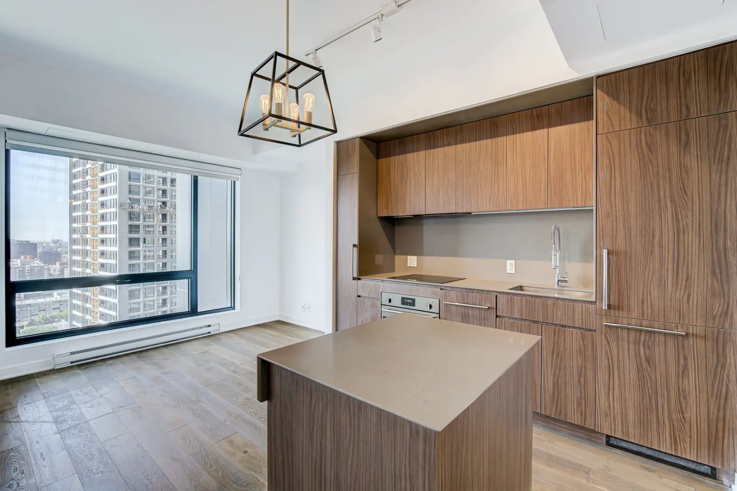 Bright modern kitchen with wood cabinets, island, and large window overlooking a city. A black geometric light fixture hangs above.