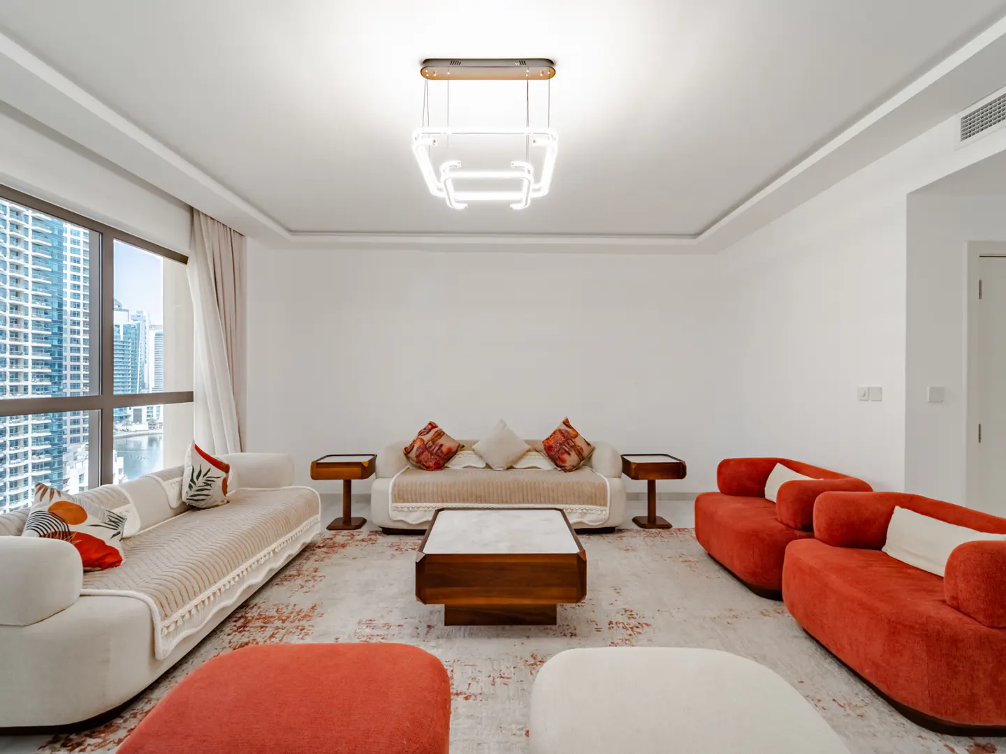 Bright living room with white walls, sofas, and a modern light fixture. A large window shows a city skyline.