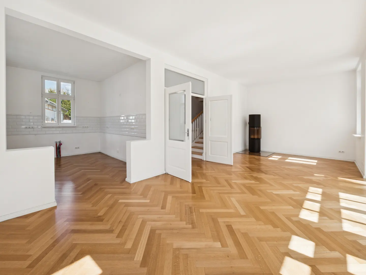 Bright, empty room with herringbone wood floors, white walls, and open doorway to a kitchen area. A black stove stands near a window.