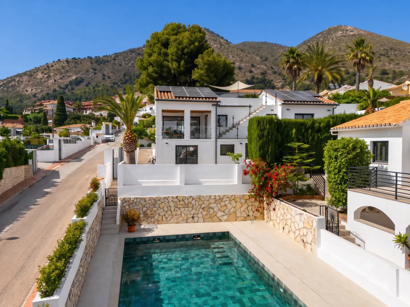 Modern white villa with a turquoise pool, stone retaining wall, and solar panels, set against a backdrop of mountains.