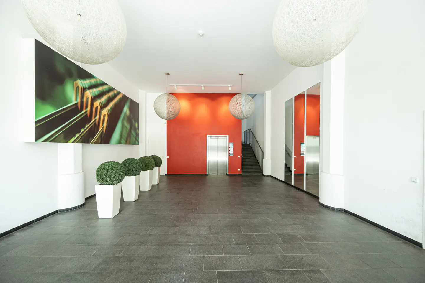 Modern building lobby with dark tile floors, a bright red accent wall, elevator, and large spherical pendant lights.