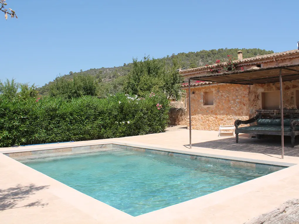 A turquoise pool sits in front of a stone house with a covered patio and green foliage.