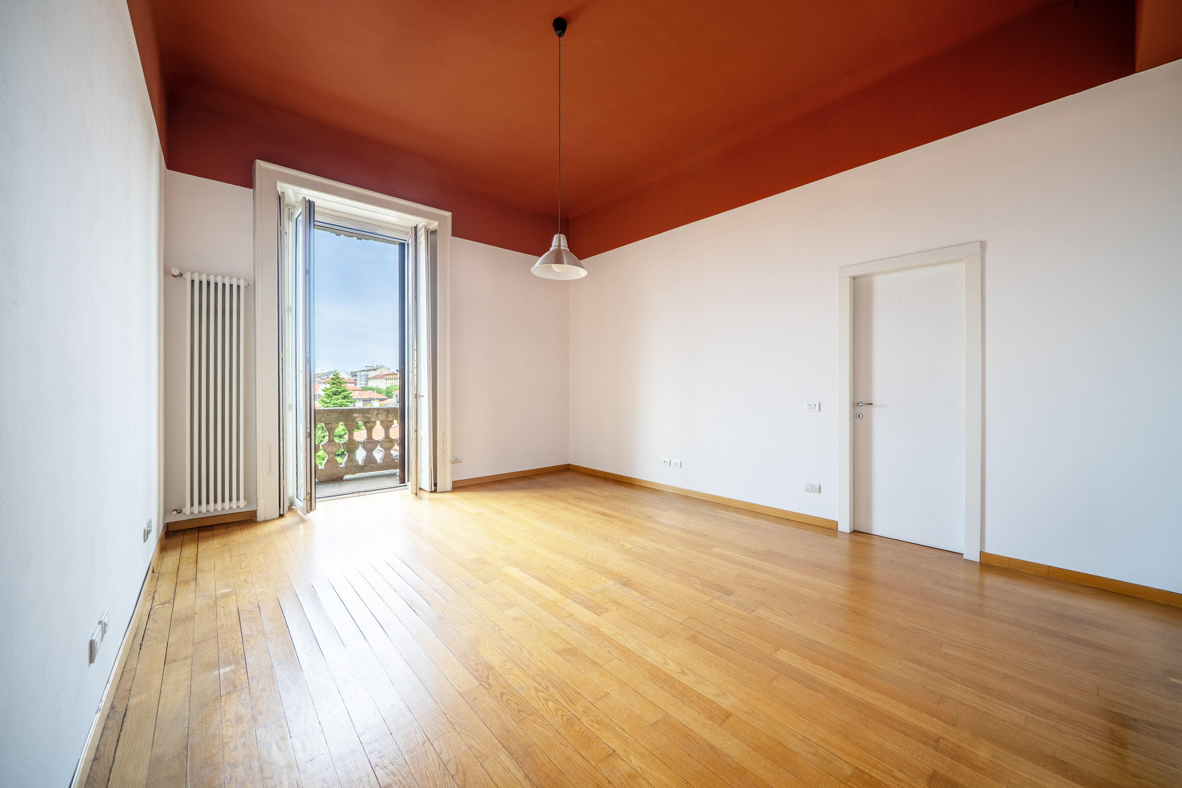 Empty room with wood floors, white walls, and a red ceiling. Balcony with open doors and a view of the city.