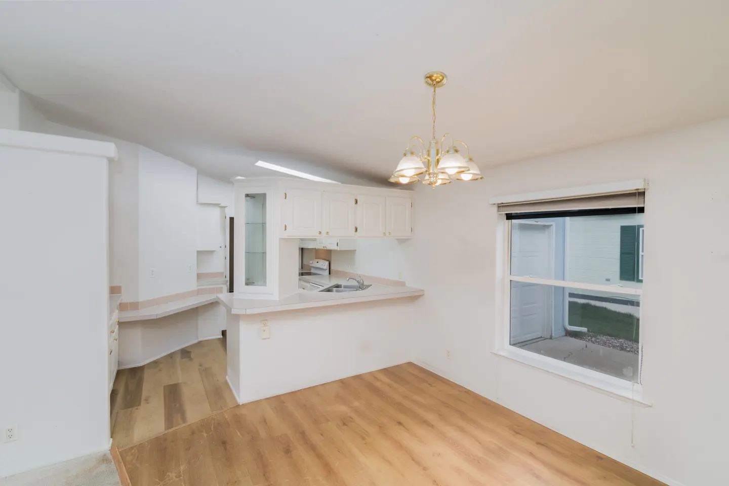 A bright, white kitchen with wood floors, cabinets, and a gold chandelier. A window looks out onto a green lawn.