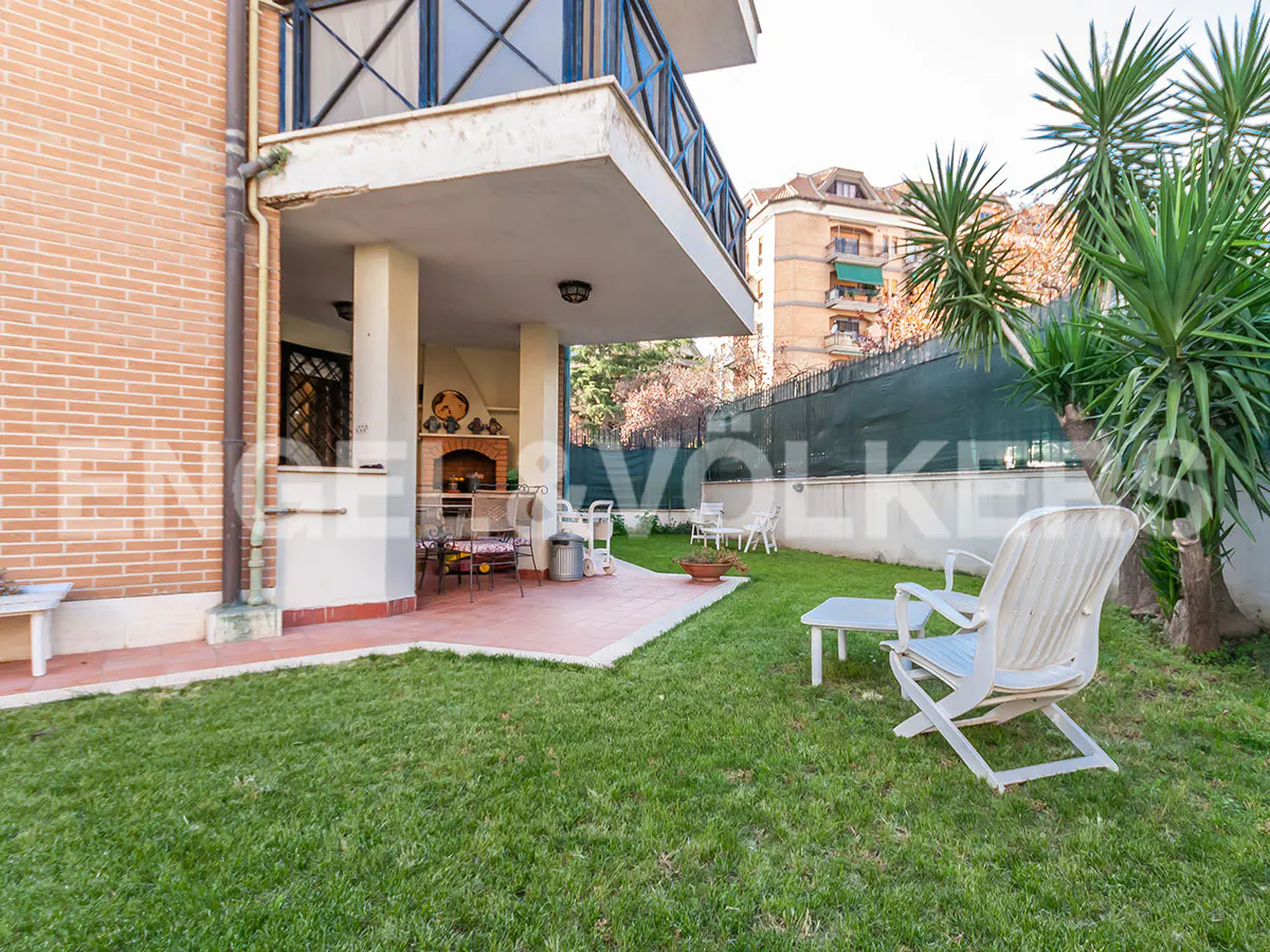 Backyard view of a brick house with a patio, grill, lawn, and white plastic chairs.