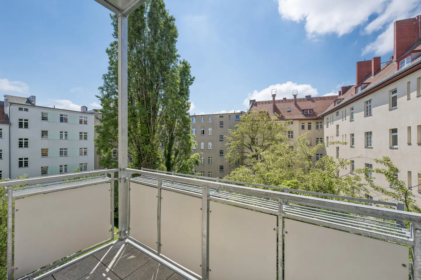 View from a balcony with metal railings and white panels, overlooking a courtyard with trees and apartment buildings.