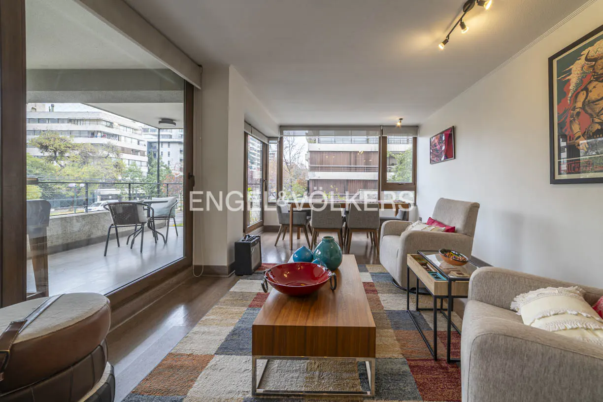 Modern living room with a wooden coffee table, beige armchairs, a colorful rug, and a balcony view of city buildings.
