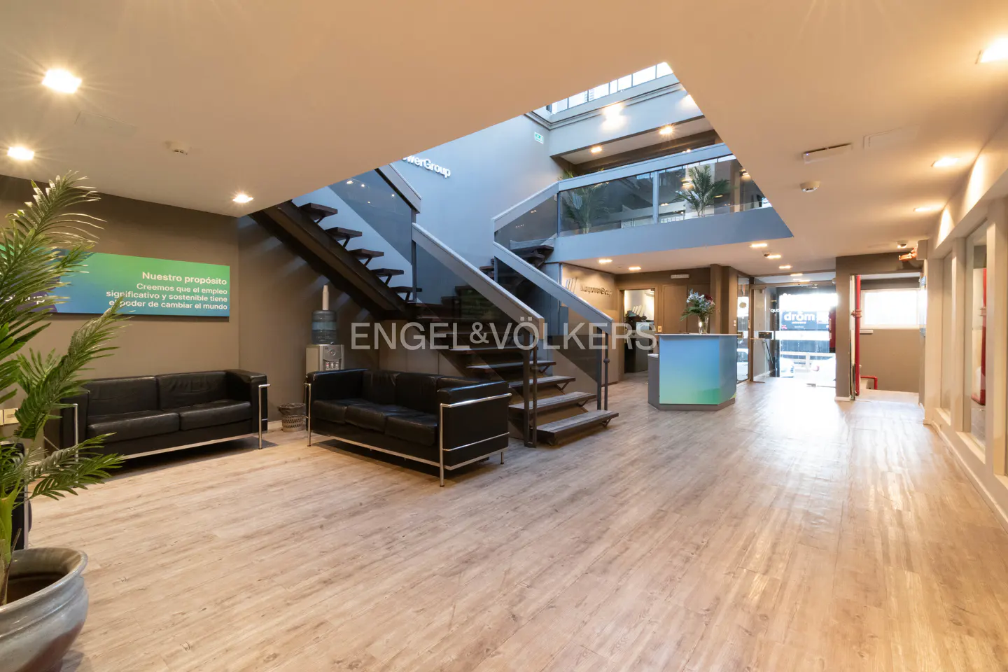 Bright real estate office lobby with black sofas, wood floors, and a staircase leading to a second floor. A reception desk is visible.