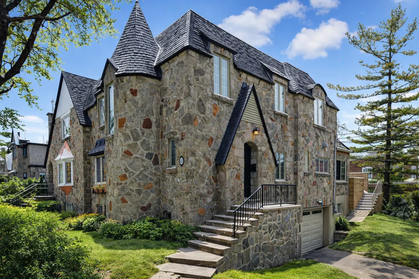 A large, two-story stone house with a turret and a dark gray roof on a sunny day.