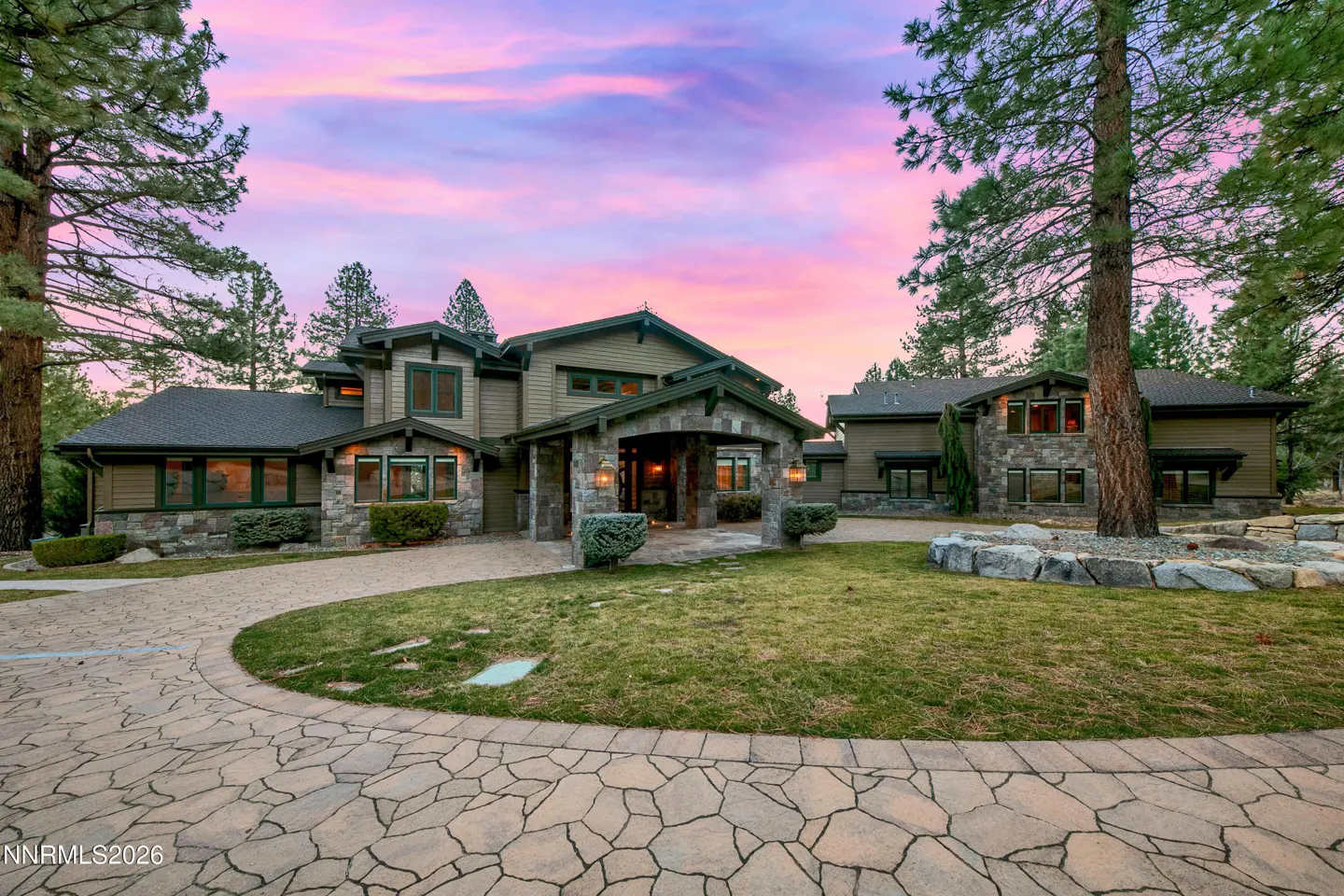 Two-story olive green house with stone accents, a stone driveway, and a pink and purple sunset sky.
