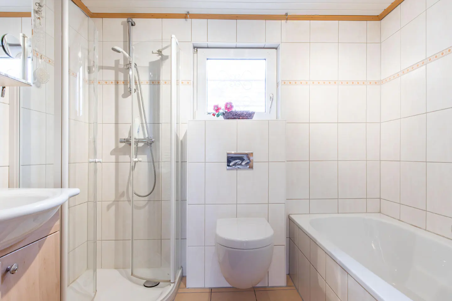 Bright bathroom with white tile walls, a shower stall, toilet, and bathtub. A small window sits above the toilet with pink flowers.