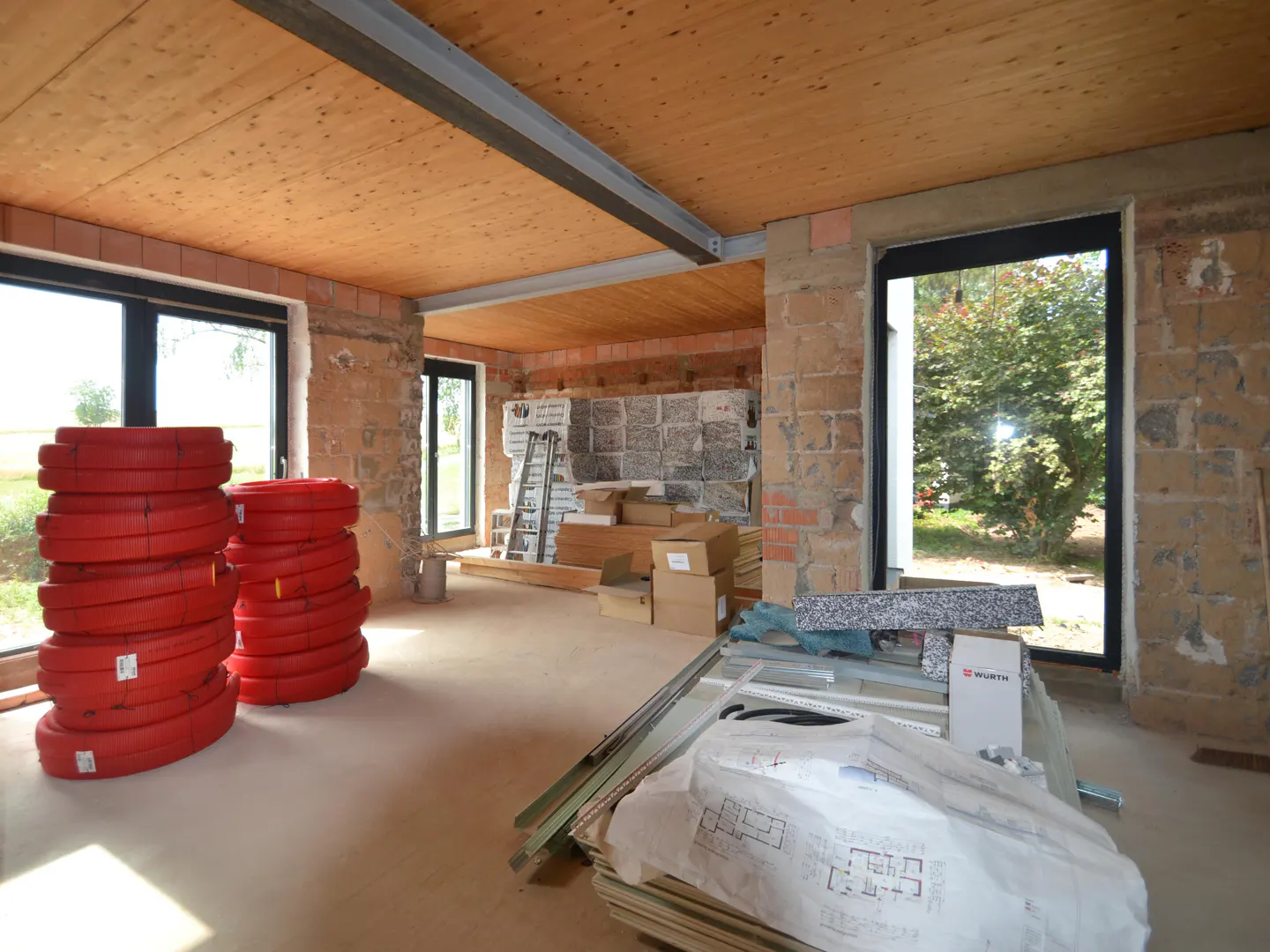 Interior view of a house under construction, showing red coils, boxes, and building plans. The walls are brick, and the ceiling is wood.