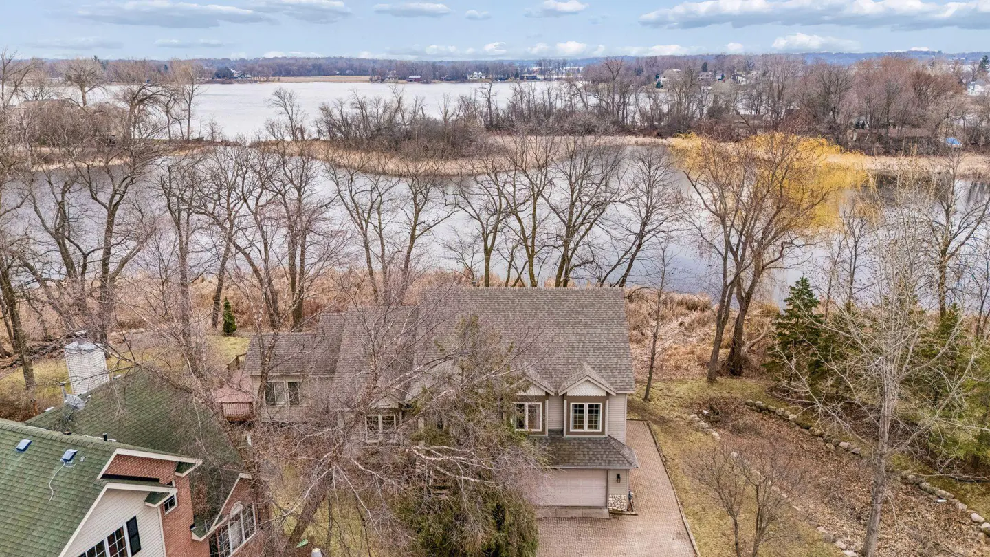 Aerial view of a gray house with a gray roof, near a lake with bare trees and a cloudy sky.
