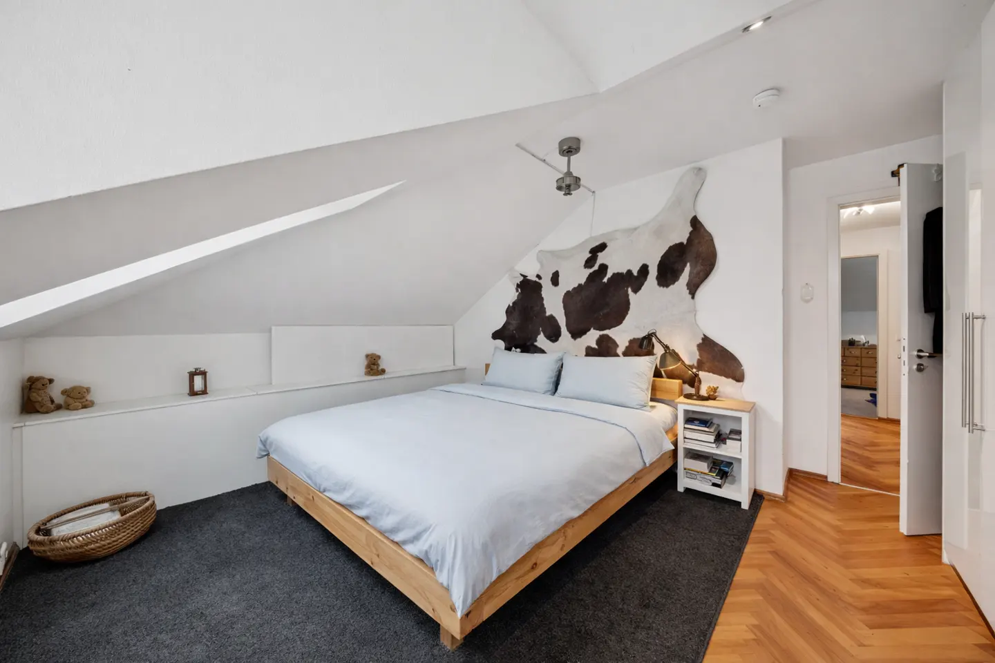 Attic bedroom with a wooden bed frame, light blue bedding, and a cowhide rug on the wall. White walls and a gray carpet.