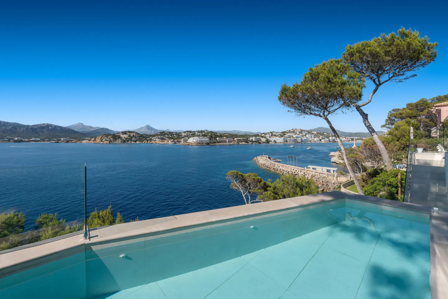 View from a pool overlooking a blue bay with mountains in the distance under a clear blue sky.