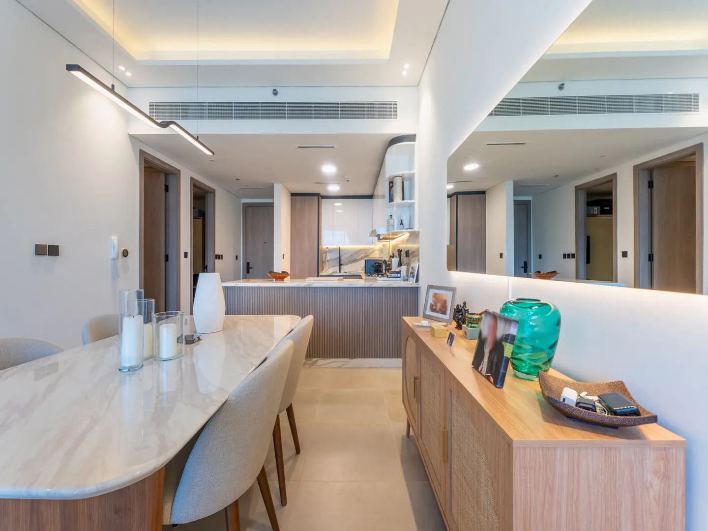 A modern apartment interior with a dining table, sideboard, and open kitchen. The color palette is neutral with white walls and light wood furniture.