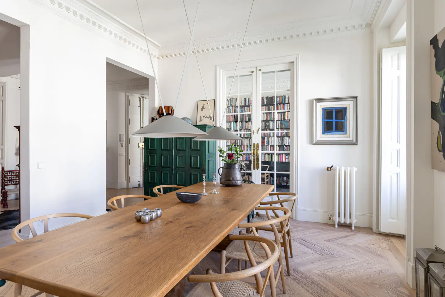 Bright dining room with a long wooden table, chairs, and pendant lights. A green cabinet and a bookcase are in the background.