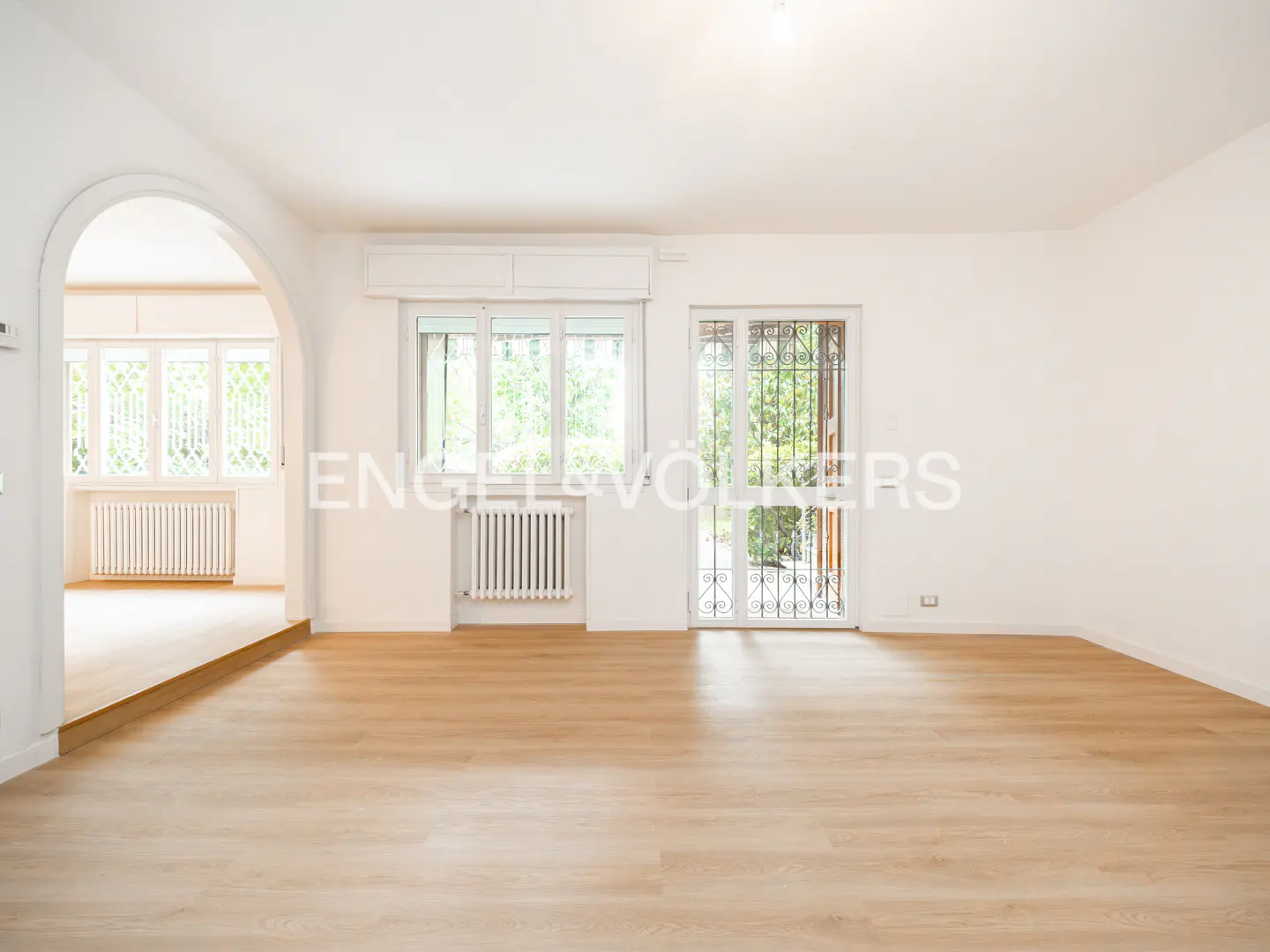Bright, empty living room with light wood floors, white walls, a large arched doorway, and windows with white radiators.