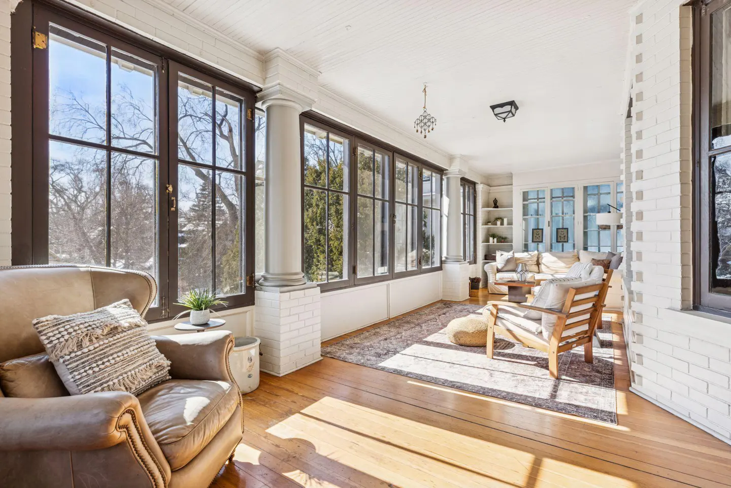 Sunroom with hardwood floors, white brick walls, and large windows. A leather chair sits near the windows.