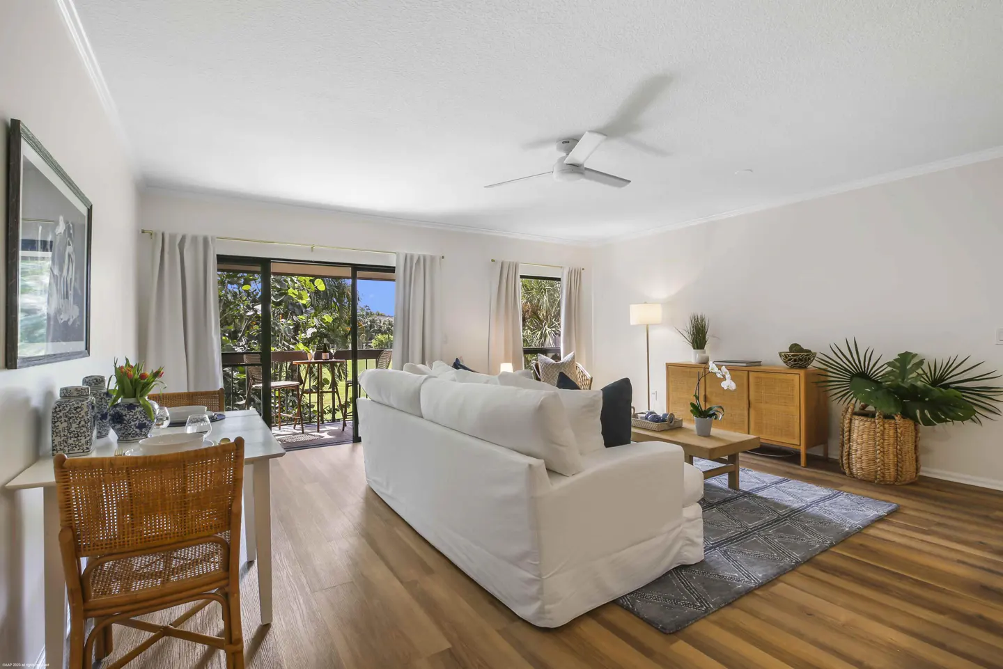 Bright living room with white sofa, wood floors, and dining area. Balcony view of lush greenery. Ceiling fan and natural light.