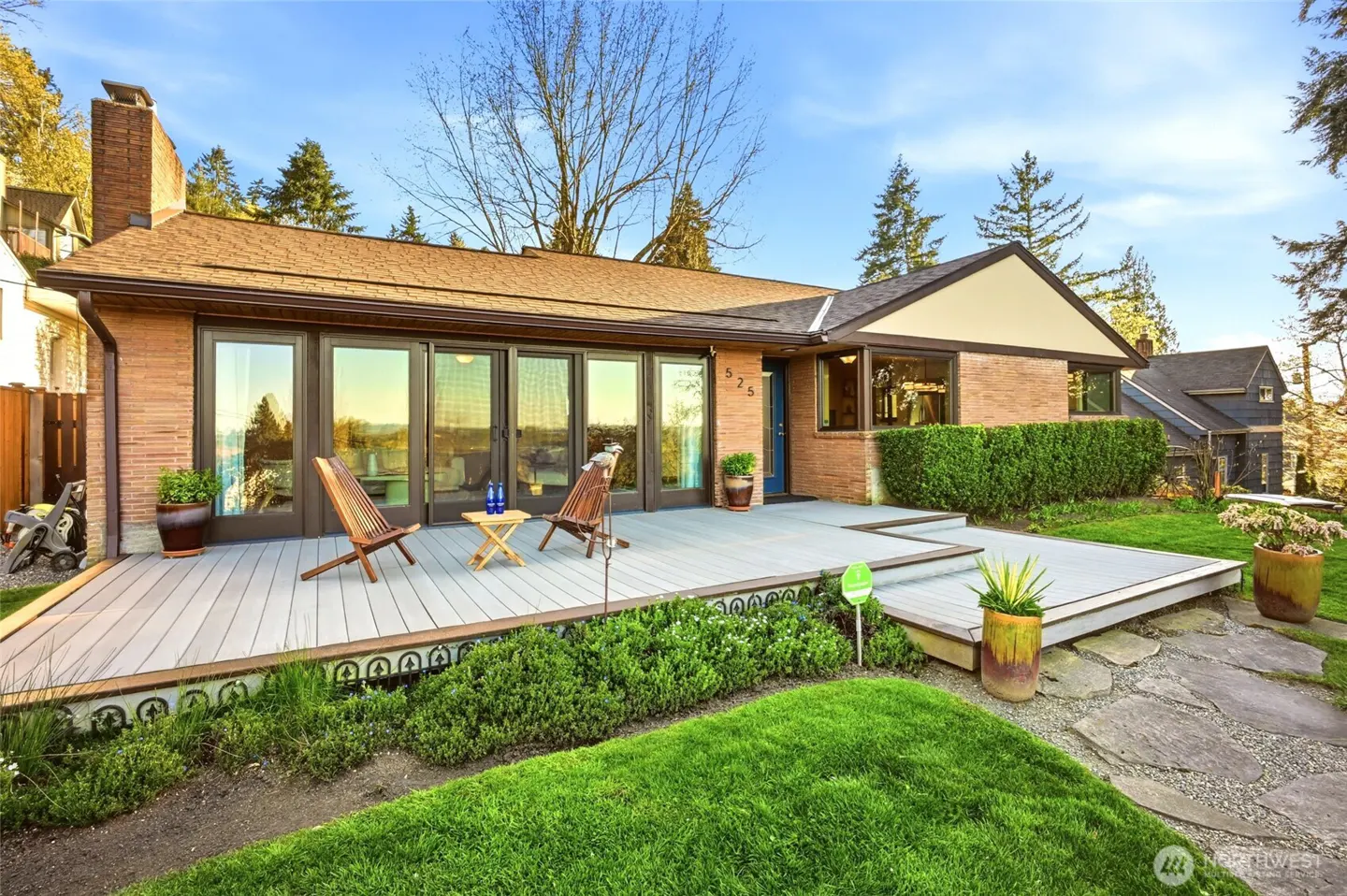 Exterior of a brick house with a brown roof, a gray deck, and a green lawn on a sunny day. Two wooden chairs sit on the deck.