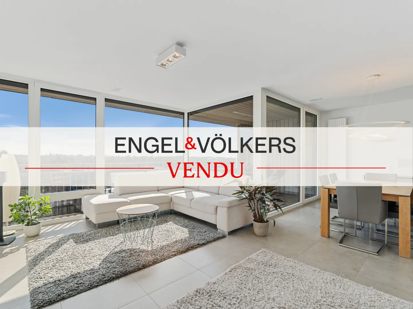 Bright, modern living room with white sofa, gray rug, and large windows. Engel & Völkers "Vendu" overlay.
