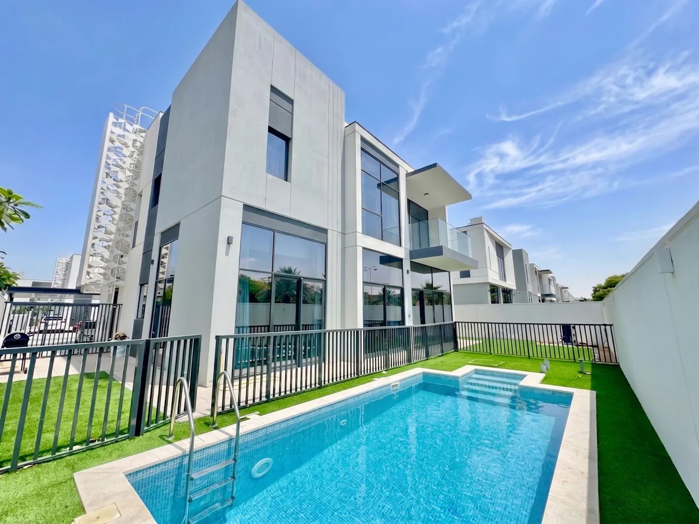 Modern white house with a blue tiled pool and green turf. Black metal fence surrounds the pool. Blue sky with light clouds above.