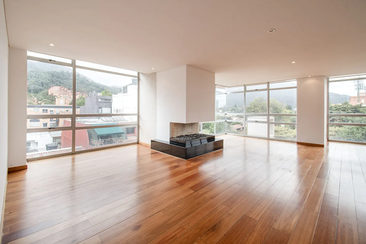 Bright, empty room with hardwood floors, a modern fireplace, and large windows with a city view.