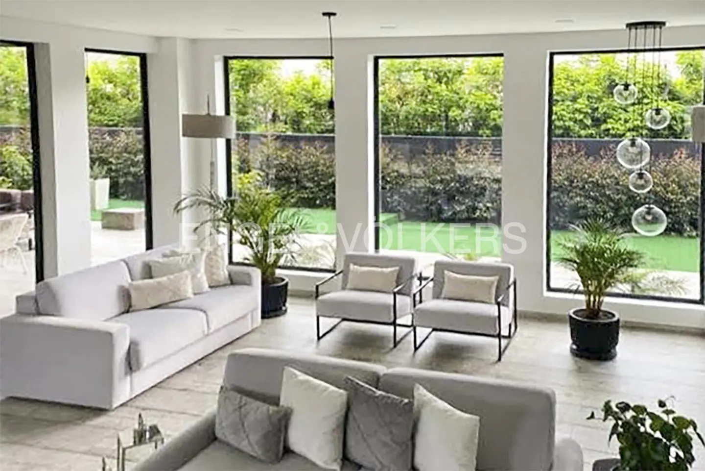 Bright living room with gray sofas, armchairs, and large windows overlooking a green garden. A modern chandelier hangs from the ceiling.