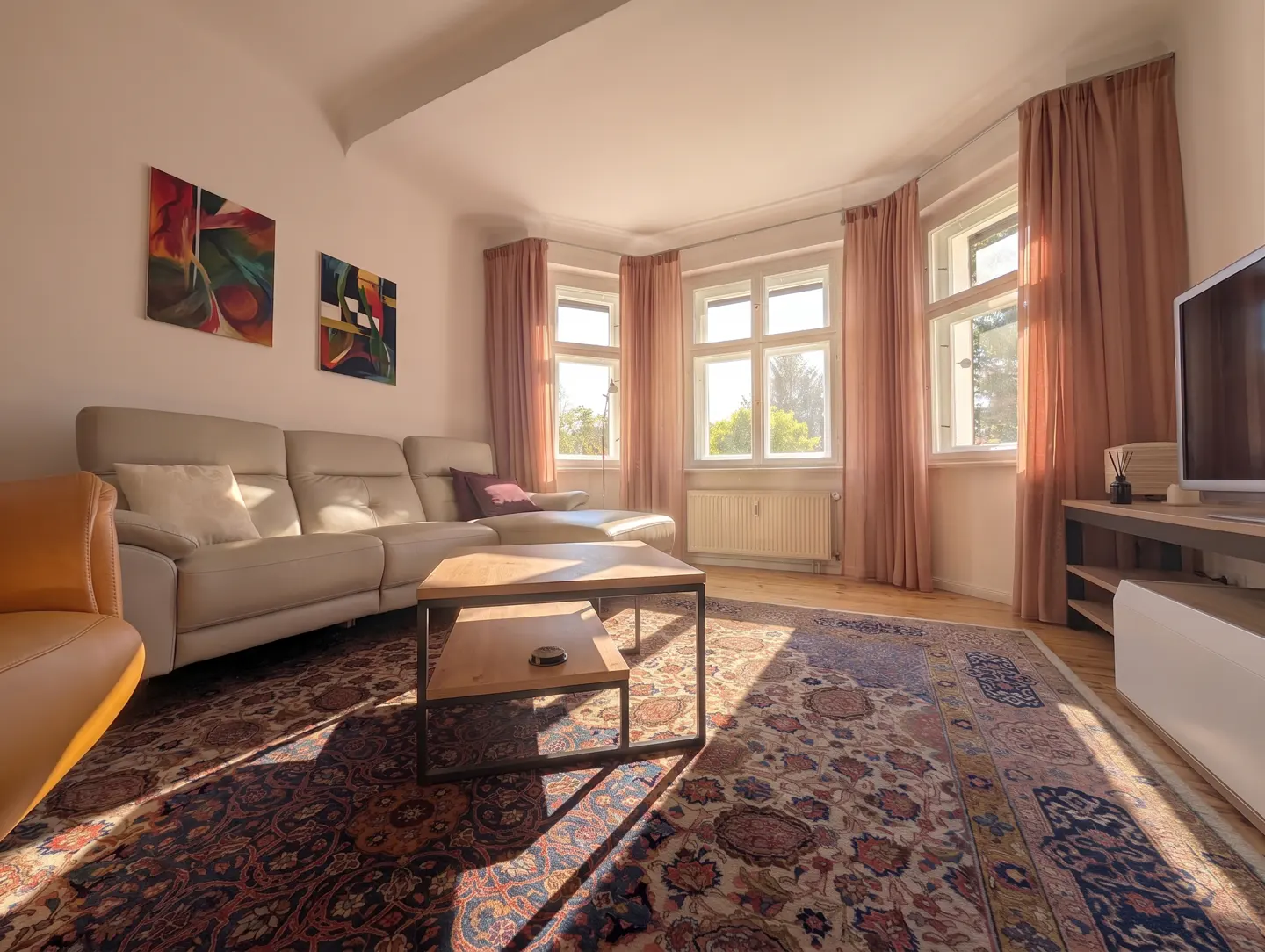 Bright living room with a beige sectional, wooden coffee table, patterned rug, and large windows with pink curtains.