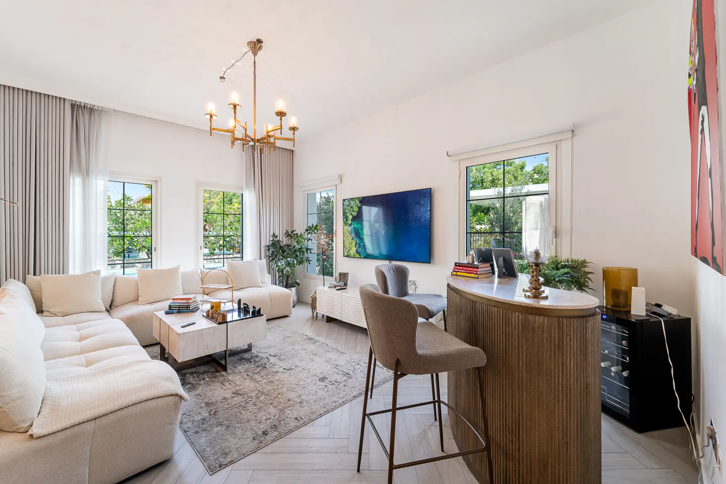 Bright living room with a white sectional sofa, marble coffee table, and a bar with two stools. A gold chandelier hangs from the ceiling.