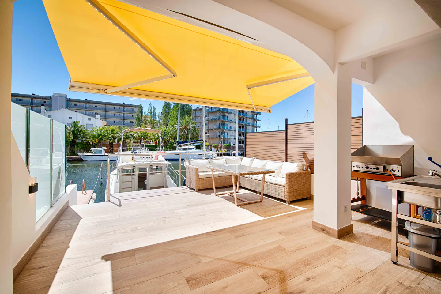 Outdoor patio with wood floors, wicker furniture, and a yellow awning overlooking a canal with boats. Grill and sink area visible.