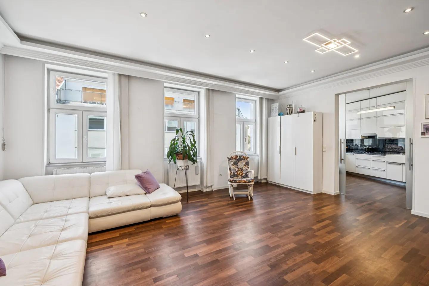 Bright living room with white sectional sofa, wooden floors, a rocking chair, and a view into a modern white kitchen.