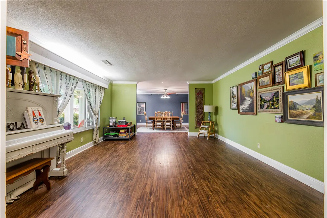 A wide shot of a living room with green walls, wood floors, and a white piano. A dining room is visible in the background.