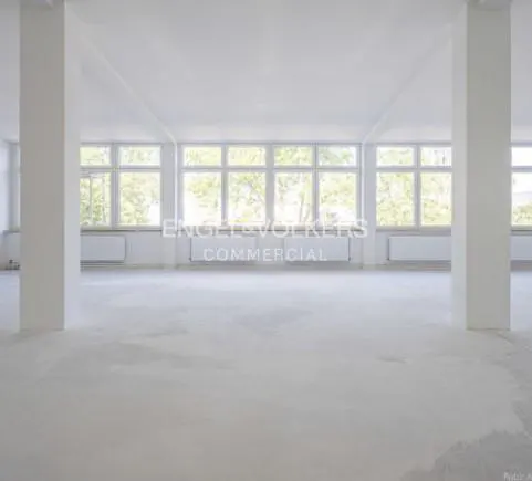 Bright, empty commercial space with white walls, pillars, and large windows. Radiators are visible under the windows. "Engel & Volkers Commercial" logo.