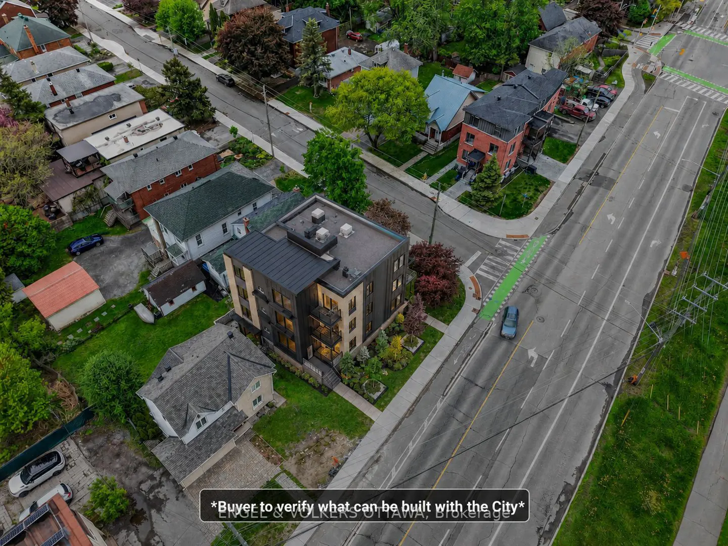 Aerial view of a modern, four-story apartment building with black and beige siding, surrounded by green lawns and trees, next to a street.