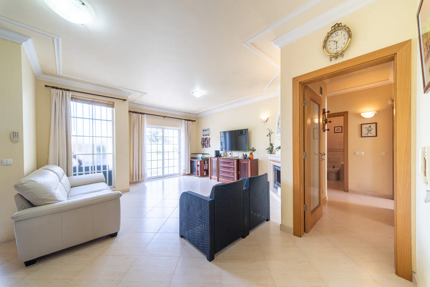 A bright living room with a cream sofa, black wicker chair, TV, and a glimpse of a bathroom through an open door.