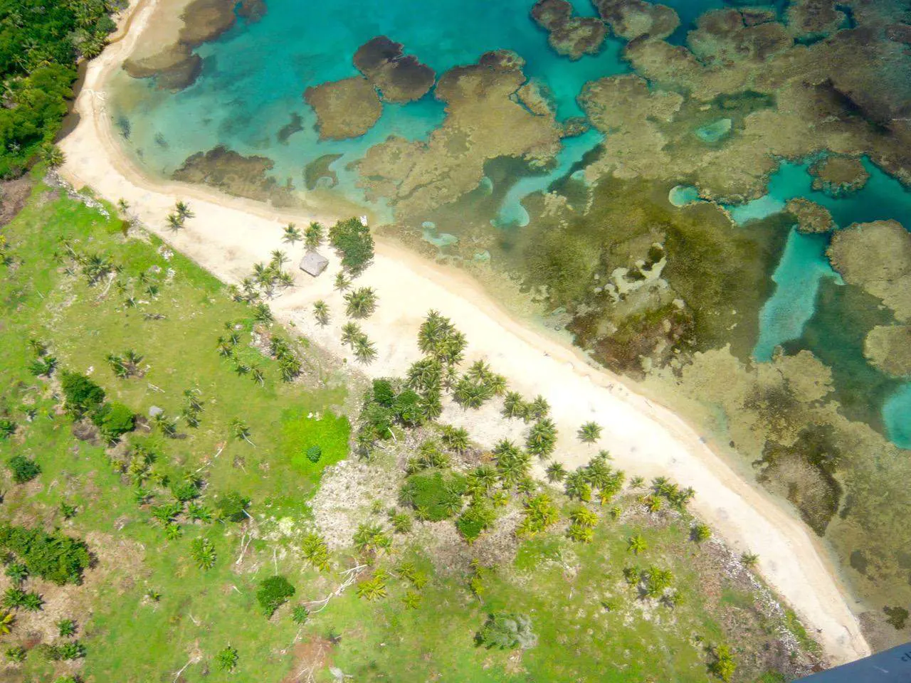 Aerial view of a tropical island with a white sand beach, turquoise water, and lush green vegetation. A small hut sits on the beach.