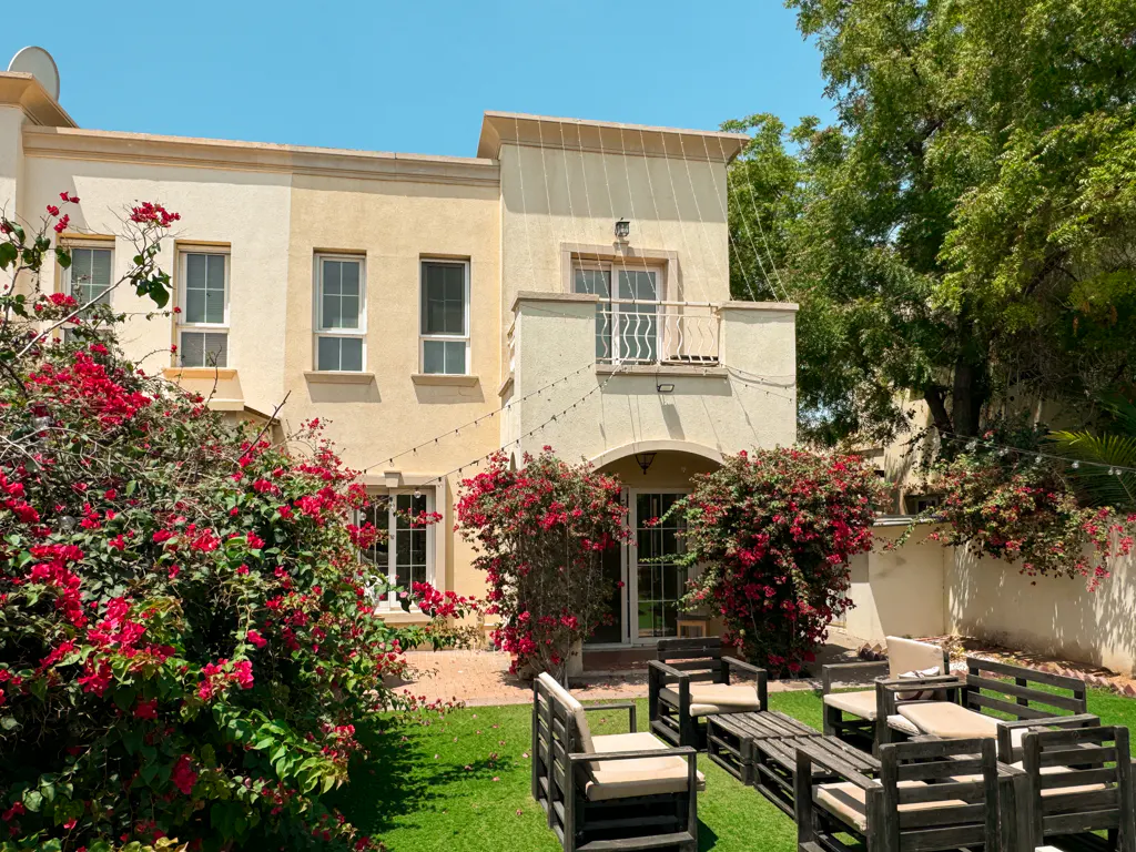 Two-story beige townhouse with a balcony, surrounded by red bougainvillea and a wooden pallet patio set on green grass.