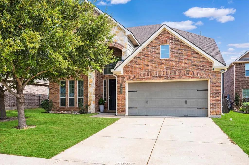 Two-story brick house with a gray garage door, a concrete driveway, and a green lawn. A large tree is on the left.