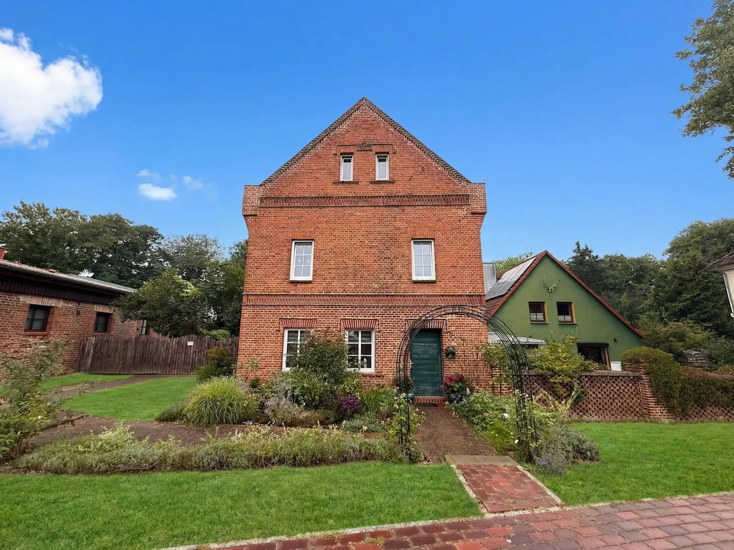 A two-story red brick house with a green door and a garden under a blue sky.