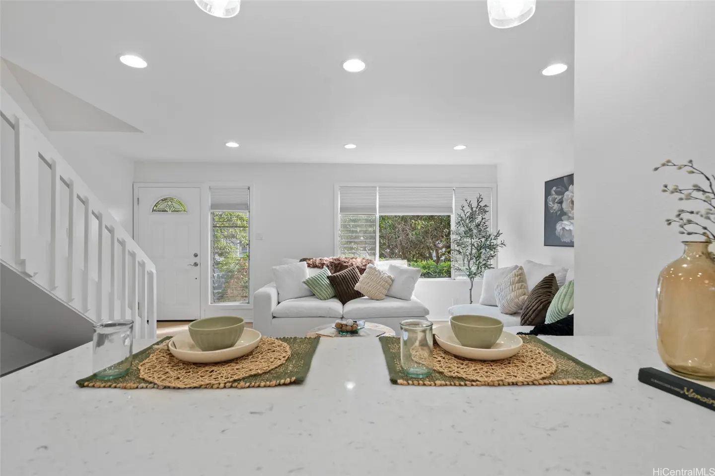 Bright, open-concept living space with white walls, a white sofa, and a kitchen island with place settings. Stairs and a front door are visible.