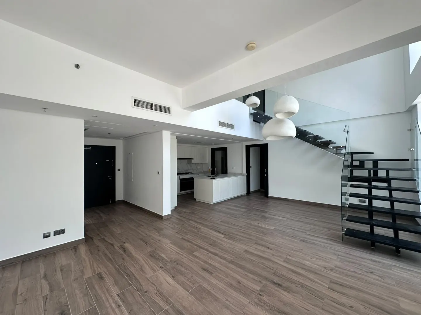 Modern open-plan living area with wood-look floors, a white kitchen island, and a black staircase with glass railings.